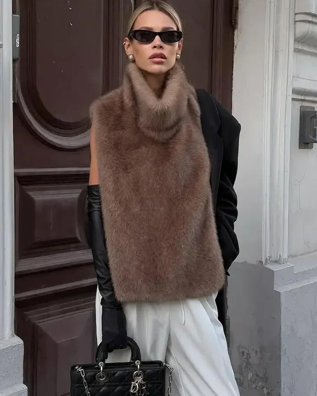 Person wearing a brown fur vest, black gloves, and sunglasses in an urban setting.
